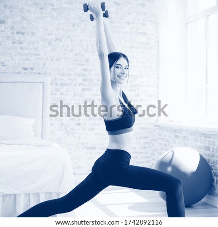 Portrait of happy smiling brunette girl in sportswear, doing fitness exercise with dumbbells, at home. Healthy lifestyle, weight lossing and sporting. Black and white monochrome blue tone picture.