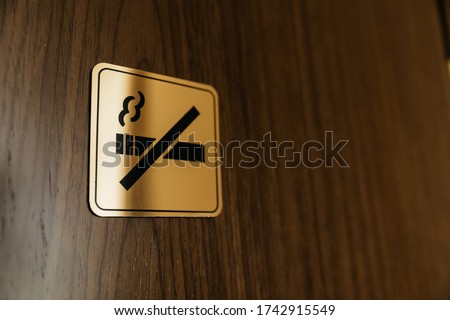 Square black and golden metallic no smoking sign on a brown wooden door inside a hotel room. Non-smoking space for incription Sign on metal texture Crossed Cigarette smoke. selective focus