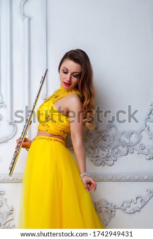 Woman in yellow dress with flute on light textured background. Flute in hand. Stylish girl with musical instrument (flute). Photo for magazine cover, website with space for an inscription or logo