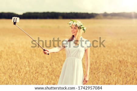 technology, summer holidays, vacation and people concept - smiling young woman in wreath of flowers taking picture by smartphone selfie stick on cereal field