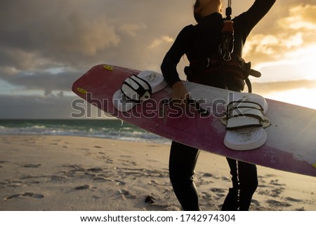 Closeup picture of professional female kitesurfer wearing a black wetsuit in kneeling position to holding up kitesurfing board equipment prior surfing sunsetting on the beach at the background
