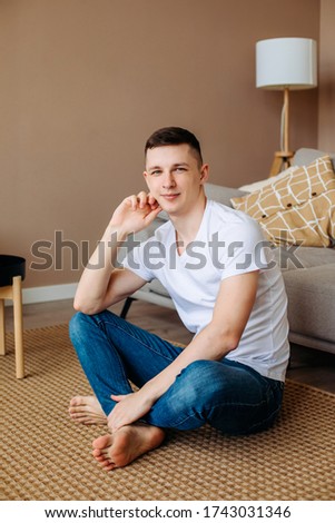 A young guy at home sitting on the floor in a beige interior barefoot and looking at the camera. Casual style
