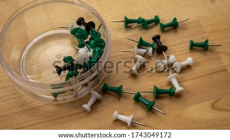Top view of full thumb tacks in a transparent box and scattered on wooden background. It is a short nail or pin used to fasten items to a wall or board for display. Selective focus on foreground.