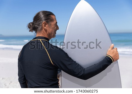Senior Caucasian man enjoying time at the beach on a sunny day, standing and holding a surfboard with sea in the background