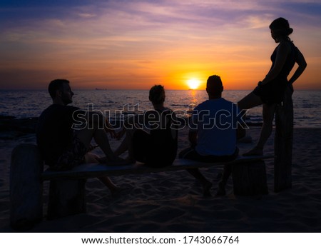Four friends enjoying the sunset on the beach. Three are sitting and one is standing on a wooden bench at the beach. In the background you can see the sun.