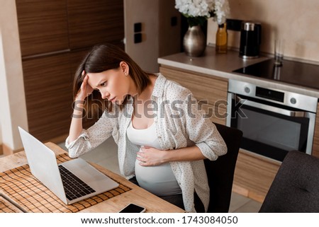 exhausted and pregnant freelancer using laptop while touching belly at home