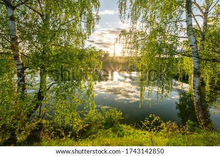 Scenic view at beautiful spring sunset on a shiny lake with green branches, birch trees, bushes, grass, golden sun rays, calm water ,deep blue cloudy sky and forest on a background, spring landscape Royalty-Free Stock Photo #1743142850