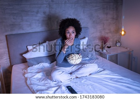 Beautiful mixed race woman sitting on bed, eating popcorn and watching a movie on TV