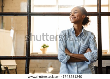 Photo of joyful african american woman with hands crossed smiling while leaning on glass wall in modern office