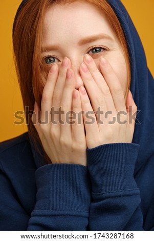 Head shot. A red-haired girl in a blue hood smiles with her eyes, covering her mouth with palms.