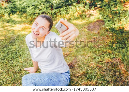Young funny girl take selfie from hands with phone sitting on green grass park or garden background. Portrait of young attractive woman making selfie photo on smartphone in summer day