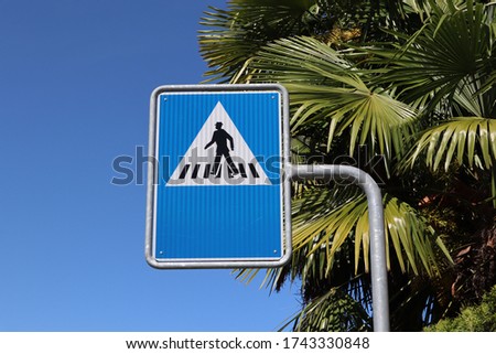 Blue rectangular sign with black and white Symbols indicating a pedestrian crosswalk in a Swiss town. There are green palm trees and blue sky in the background.