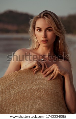 portrait of a blond girl covered herself with a big straw summer hat on the beach and looking at camera