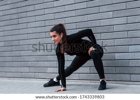 Young sportive girl in black sportswear outdoors doing stretching near gray wall.