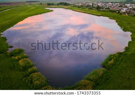 A small picturesque lake among green fields with reflection in it of beautiful clouds of the sunset sky. Aerial view