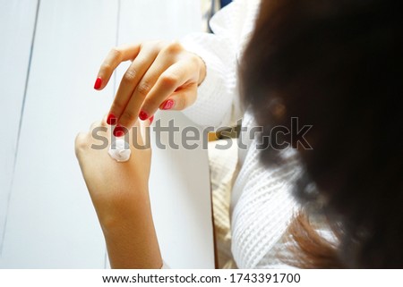 Beauty and body care theme young woman in white bathrobe applying cream on her backhanded. Beautiful female hand and fingers with red nail polish taking care of her dry skin by applying cream.