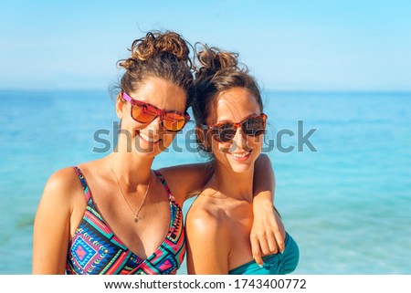 Two caucasian women sisters or friends girls in bikini on the beach at seaside in summer day on the vacation smiling wearing sunglasses in front of the sea front view