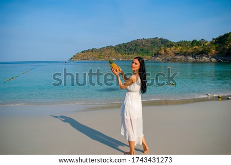 A romantic young brunette women in a long white dress holds fruit pineapple in her hands. A resting tourist on the sea. Tropical Andamand sea beach and palm trees Southeast Asia, Thailand.