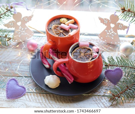 Two cups with a drink, marshmallows, caramel sticks, Christmas decor, illuminations on a wooden surface on a window background