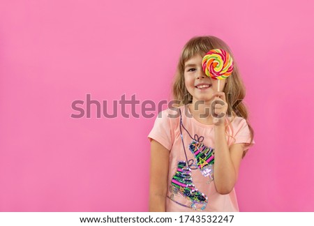 A little girl is looking through a lollipop. Little girl on a pink background. Happy child with candy in hands.A little girl holds in her hands a multi-colored candy on a stick.Fashion look.candy shop