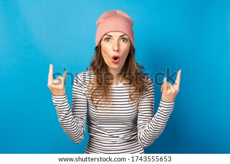Cute young woman in a hat and striped sweatshirt makes eating rock and roll with her hands on a blue background. Emotional face. Gesture party, in the gap, rock
