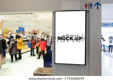 Mock up blank vertical LED board with clipping path in black frame on wall of shopping mall, blurred people wearing winter coat, empty space for insert advertising or announcement information