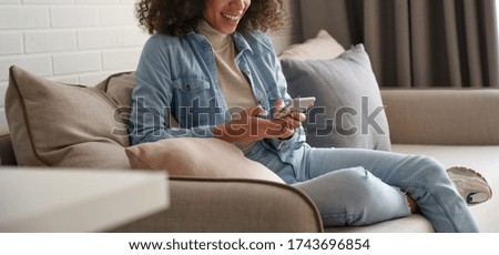 Smiling afro american millennial woman user holding smart phone watching video, playing game, shopping, chatting in mobile technology application sitting on sofa at home. Close up view, banner design. Royalty-Free Stock Photo #1743696854