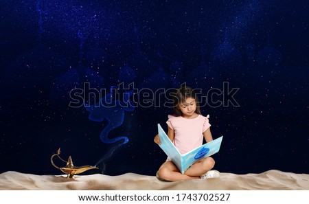 Cute little girl reading magic book and Aladdin magic lamp on sand at night