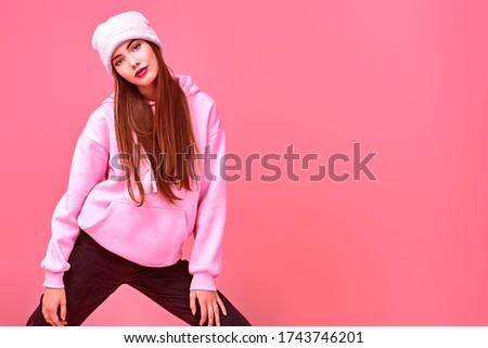 Youth fashion. Modern teen girl in a pink sweatshirt and a hat posing at studio on a pink background. Cosmetics and make-up. Copy space.