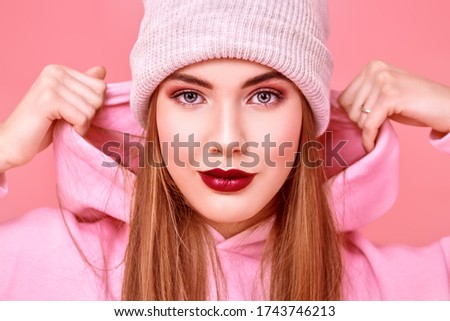 Youth fashion. Modern teen girl in a pink sweatshirt and a hat posing at studio on a pink background. Cosmetics and make-up.