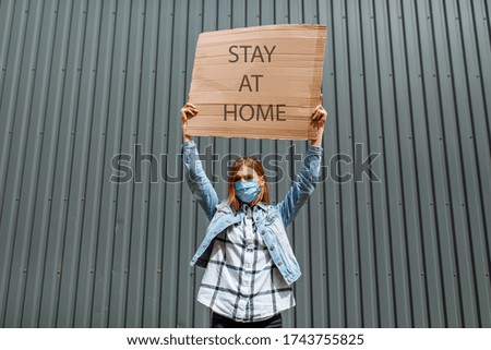 A young woman in a medical protective mask holds a cardboard placard with the words Stay at home, standing in the city against a gray wall. Quarantine, coronavirus