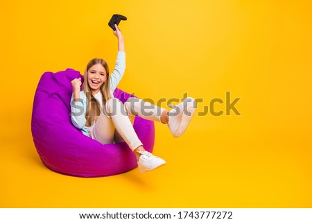 Full body photo of beautiful little lady good mood playing video games joystick sitting cozy violet bean bag rejoicing win wear casual shirt trousers shoes isolated yellow color background