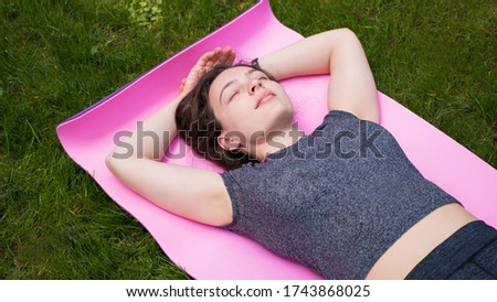 Young sporty woman in grey top lying with eyes closed in shavasana,corpse posture after yoga, workout training exercises,resting after practice, meditating,breathing deeply on pink mat on green  Royalty-Free Stock Photo #1743868025