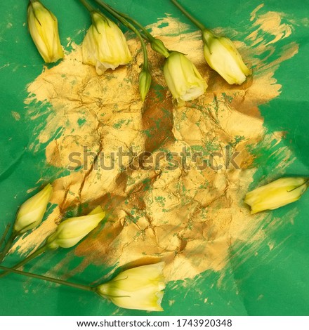 Yellow tiny rose eustoma flower buds on crumpled paper with gold gouache acrylic paint strokes on green background. An abstract background backdrop for postcards with free blank copy space for text.