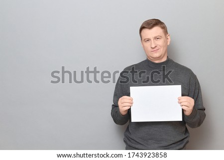 Studio portrait of proud confident blond mature man wearing jumper, holding white blank paper sheet with place for your text in hands, standing over gray background, copy space on left