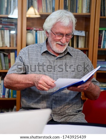 Portrait of a white bearded senior man with grey shirt reading a blue notebook, book while sitting in front of a huge book shelf in the background.                             