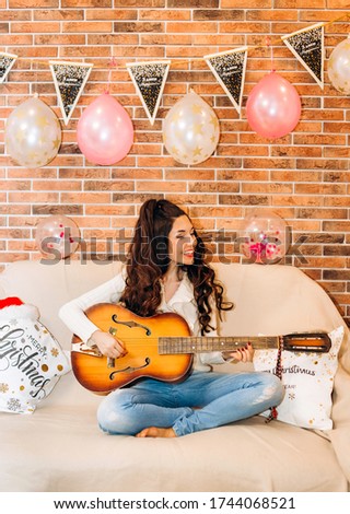 Stylish girl with a beautiful hairstyle sits on the bed and plays the guitar on a holiday day. The inscription on the triangular cards: "Happy Birthday"