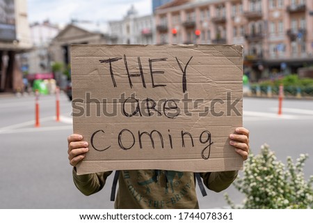 suspicious and warning inscription on sign. They are coming. Hands holding banner outside on city streets. social protest motivation concept. Guy with sign imitation. Aliens guests waiting invitation Royalty-Free Stock Photo #1744078361