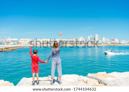 Mother with cheerful young son waving hand stare at the sea on light blue sky sun shining outdoors background. Amazing happy summer travel holiday, beauty freedom. Back view picture