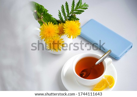 Morning tea with sweet lemon candy and light blue mobile phone, bouquet of yellow spring dandelion flowers. Beautifully styled feminime elegant white porcelain tea scene.