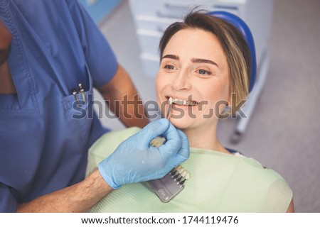 Dentist checks the level of patient's teeth whitening with a dentist's color. Dental equipment in dentistry office. Stomatology concept. Doctor's hands in medical gloves and smiling happy woman.