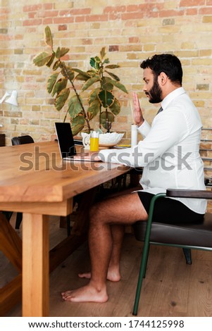 bearded african american man in shirt and underwear sitting near laptop and waving hand while having video call