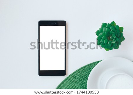 Smartphone lying on white table, cup of cappuccino and green plant. Top view with copy space