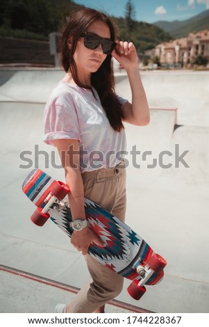 Attractive young woman with colorful skateboard dressed in casual t-shirt and trousers standing on the white skate ramp in the park with beautiful city view, green mountains and blue sky backward