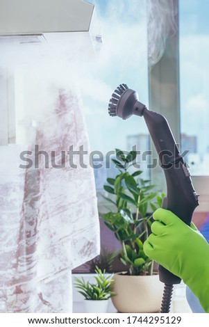 House cleaning. The girl holds an electric steam cleaner, which cleans the kitchen extract, the surface, with hot wet steam. Cleaning of a surface. Green gloves. Light apartment. closeup.