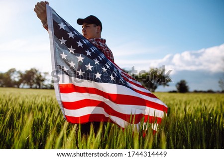 Closeup of a young patriotic farmer stands among new harvest. Boy walking with the american flag on the green wheat field celebrating national independence day. 4th of July concept.