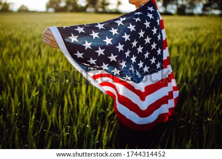Closeup of a young patriotic farmer stands among new harvest. Boy walking with the american flag on the green wheat field celebrating national independence day. 4th of July concept.