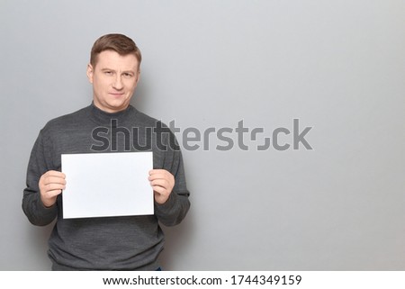 Studio portrait of happy blond mature man wearing jumper, smiling cheerfully, holding white blank paper sheet with place for your text in hands, standing over gray background, copy space on right
