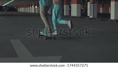 Smiling woman standing on a longboard while her friend is pushing her behind and running in car parking. Enjoying life. Urban city lifestyle