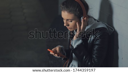 Young skater happy teen girl using a smart phone. Young caucasian woman sitting on skateboard listening to music on smartphone late at night in dark underground tunnel